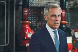 Prime Minister Mark Carney in a dark suit and blue tie, photographed in front of industrial pipefitting equipment at a Calgary union training facility.